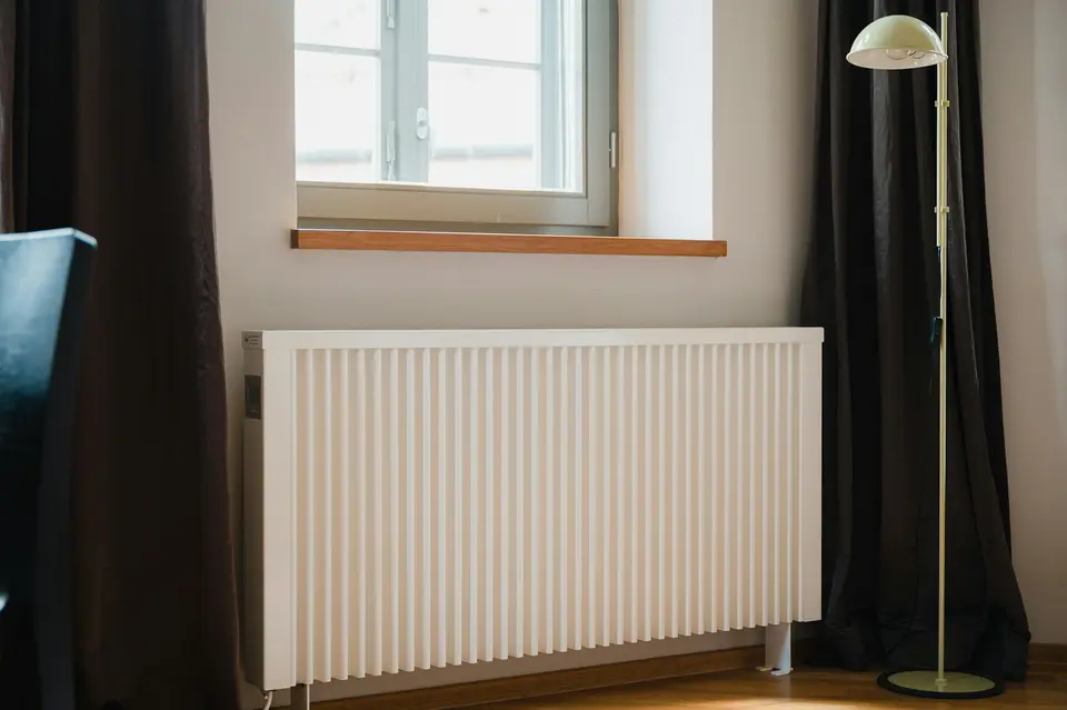 Witte paneelverwarming onder een raam, met donkere gordijnen en een staande lamp
